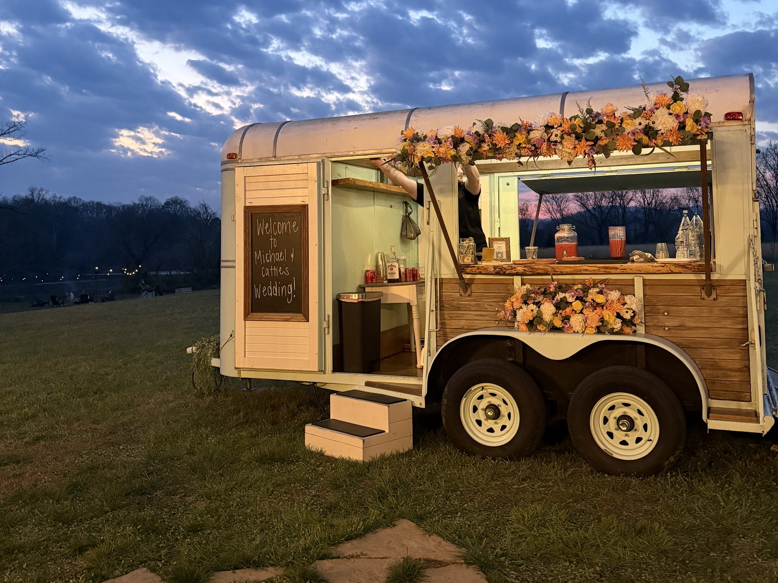 Moon & Rae's vintage horse-trailer mobile bar, lit up at dusk with a glowing BAR sign and string lights